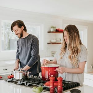 Happy couple cooking at the kitchen island bench surrounded by red le creuset dishes